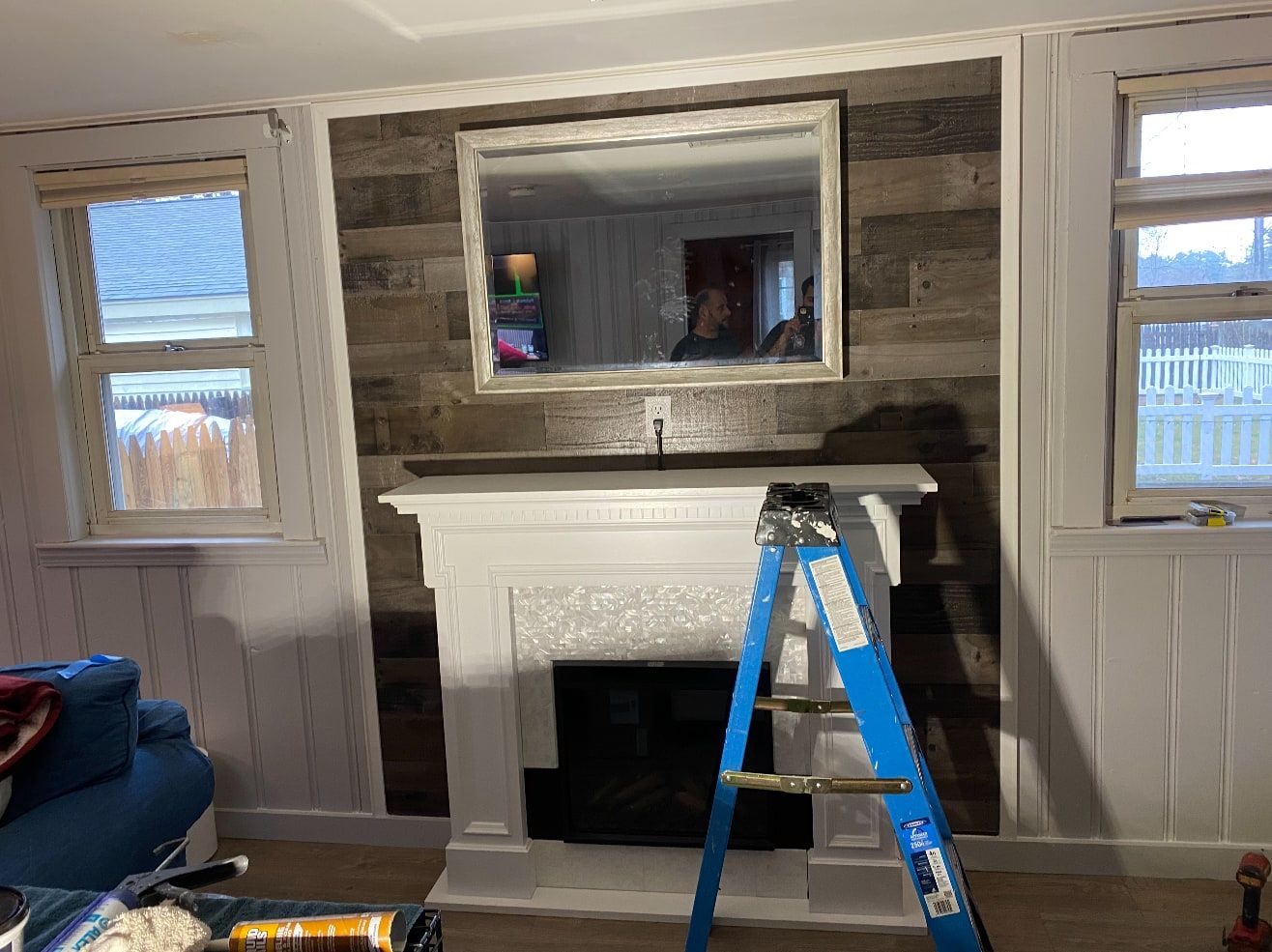White fireplace mantel surround set against a rustic barn-wood accent wall with a mirror above, between two windows