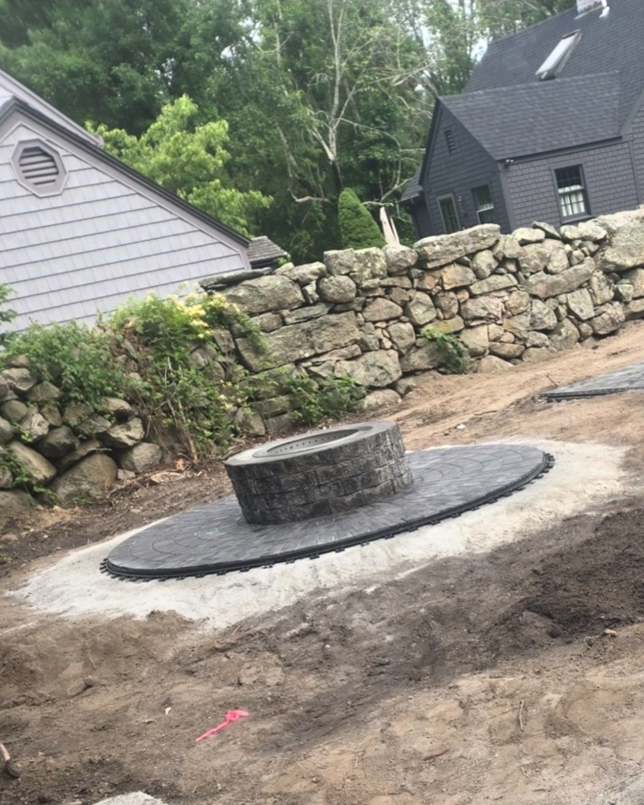 New stone fire pit on a round dark paver pad set in fresh base material, in front of an old fieldstone wall