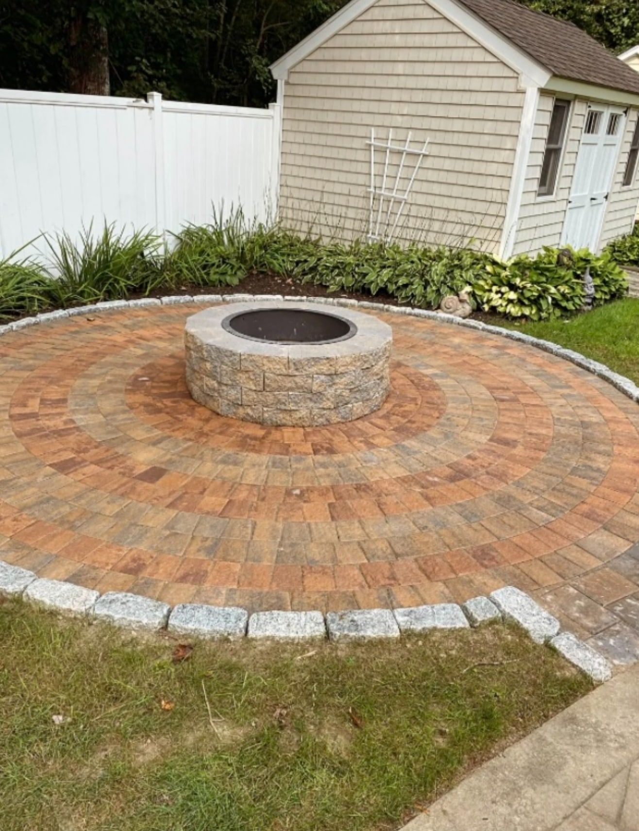Circular paver patio in warm red and tan tones with a stone fire pit at the centre and granite cobble edging, beside a shed and planted border