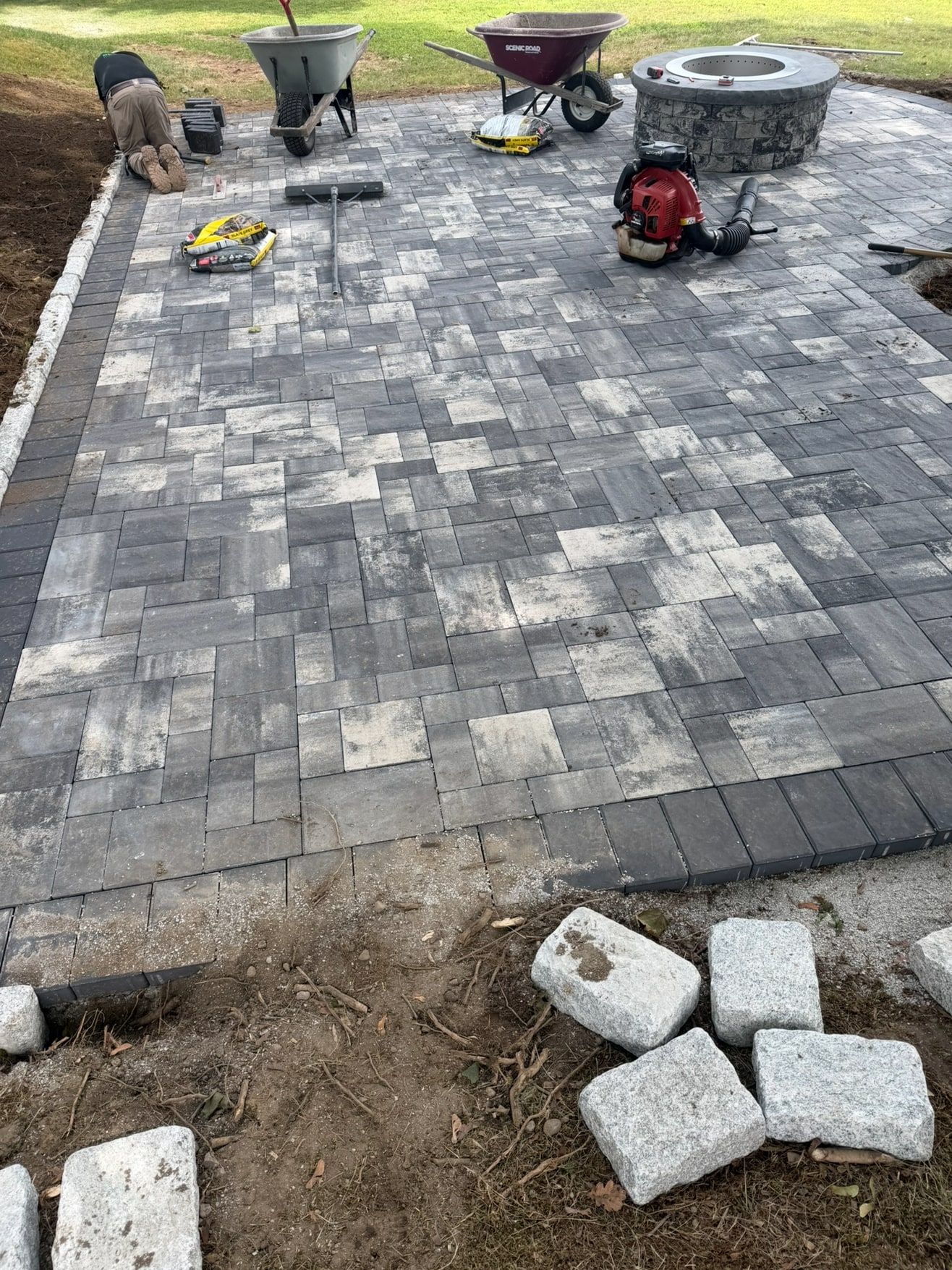 Large grey blend paver patio being installed around a fire pit, with granite edging stones ready to set