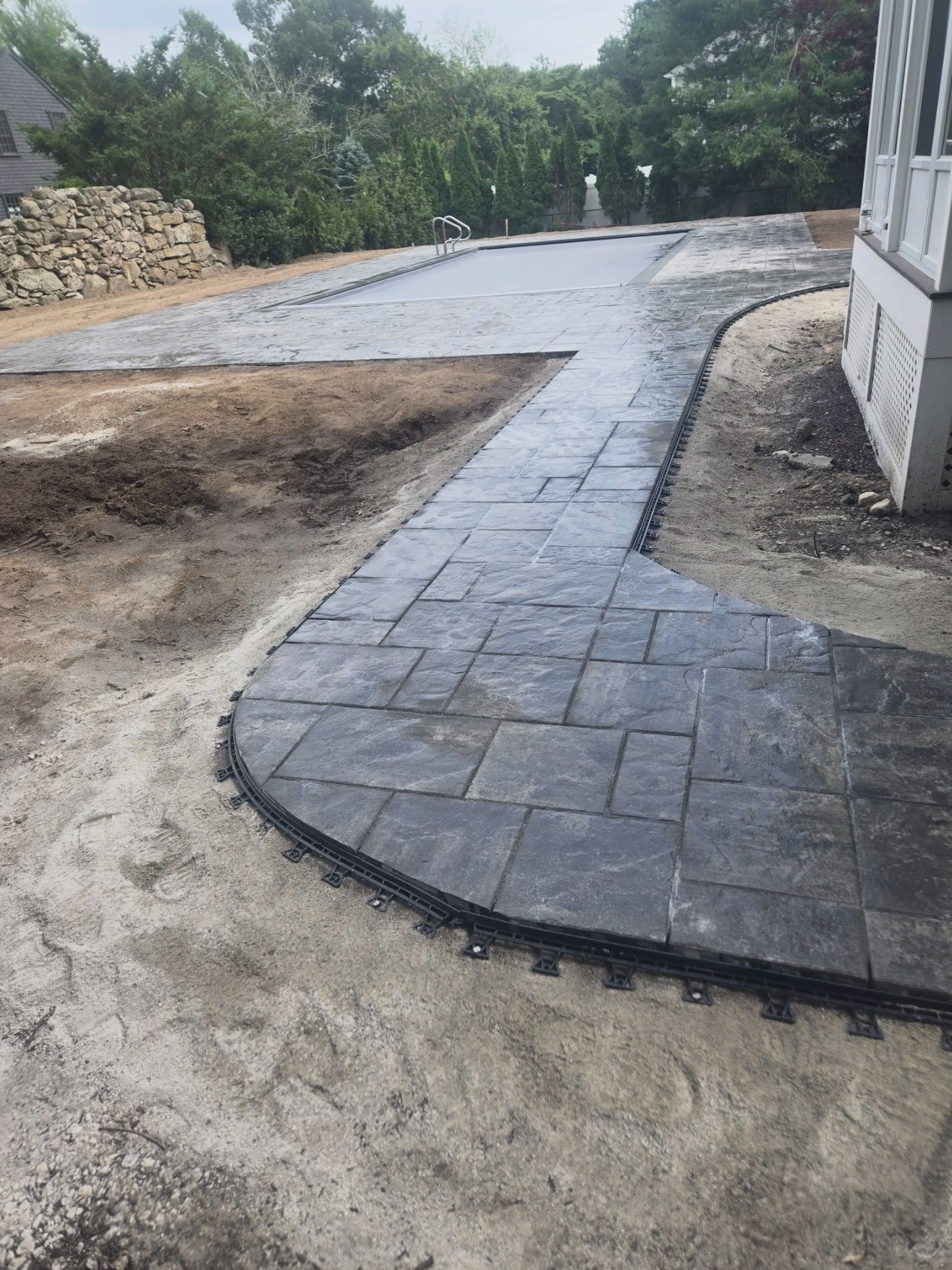Curved dark-grey paver walkway leading from the house to a large paver pool patio, with edge restraint along the border