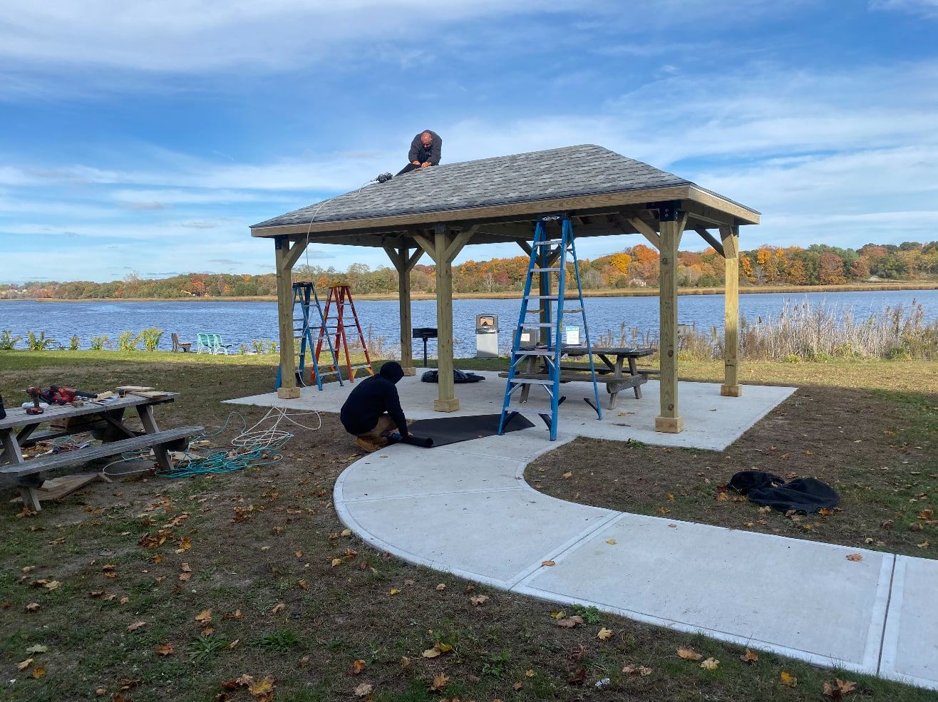 Timber-frame pavilion with a shingled hip roof being finished on a concrete pad beside the water, with ladders and crew on site